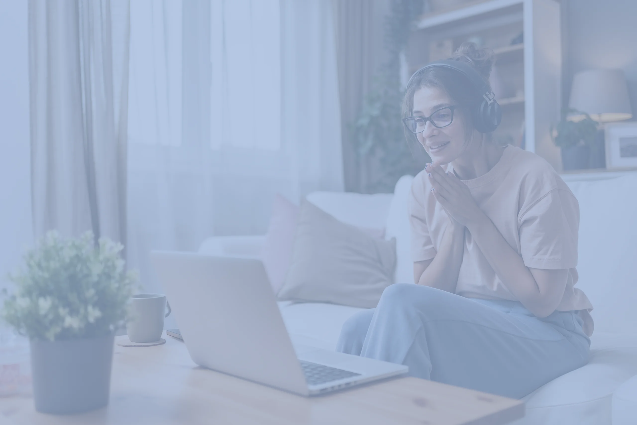 Online behavioral health. A woman wearing headphones and glasses sits on a sofa, smiling at a laptop. A cup and plant on the table, soft natural light through curtains.