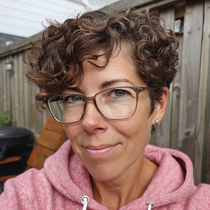 A person with short curly brown hair, glasses, and a pink hoodie smiles slightly while sitting outside near a wooden fence.