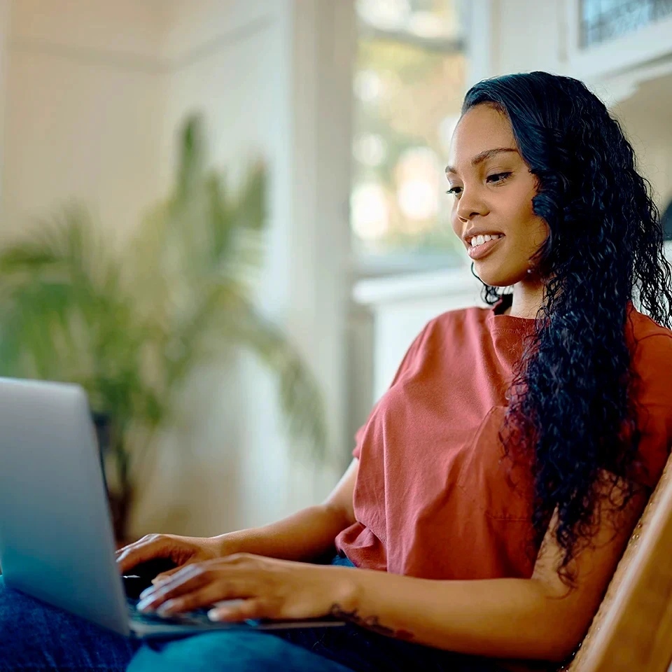 A woman with long curly hair, wearing a red t-shirt and jeans, sits indoors using a laptop. She is smiling and appears focused, with a blurred plant and window in the background.