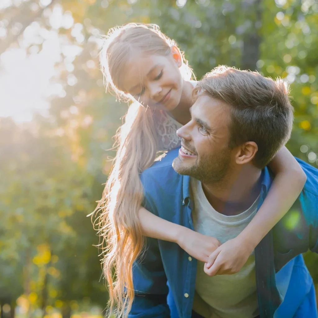 A smiling man gives a piggyback ride to a young girl outdoors, both looking at each other happily with sunlight streaming through green trees in the background.