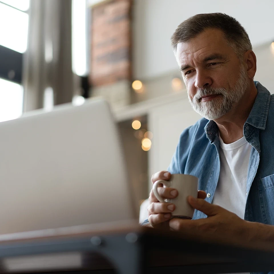 A middle-aged man with gray hair and a beard sits at a table, holding a white mug and looking at a laptop screen. He is wearing a denim shirt over a white t-shirt in a cozy, well-lit room.