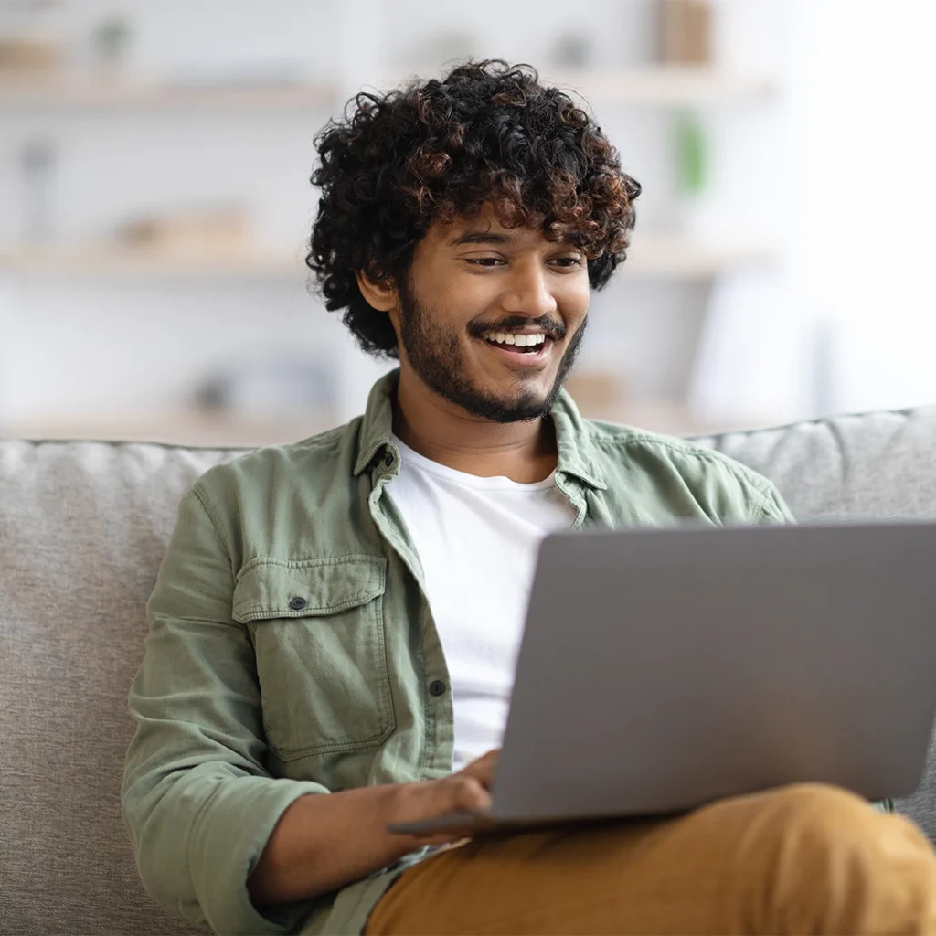 A young man with curly hair, wearing a green shirt and white t-shirt, sits on a couch smiling while using a laptop. The background is softly blurred with shelves and decor visible.