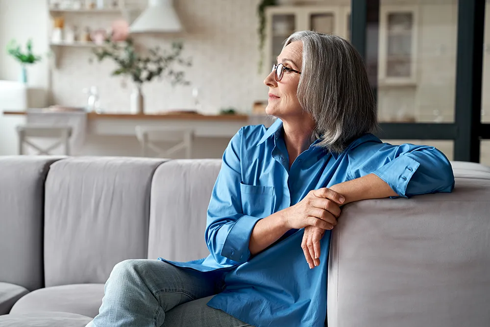 Older woman with gray hair and glasses, wearing a blue shirt and jeans, sits on a light gray couch in a bright living room, looking thoughtfully out of frame to her left.