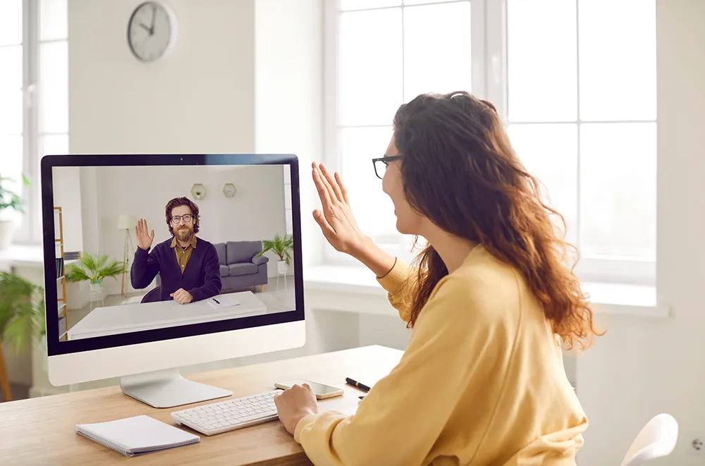 A woman sits at a desk and waves at a man on a computer screen during a video call. Both are smiling and appear to be in bright, well-lit rooms with large windows.