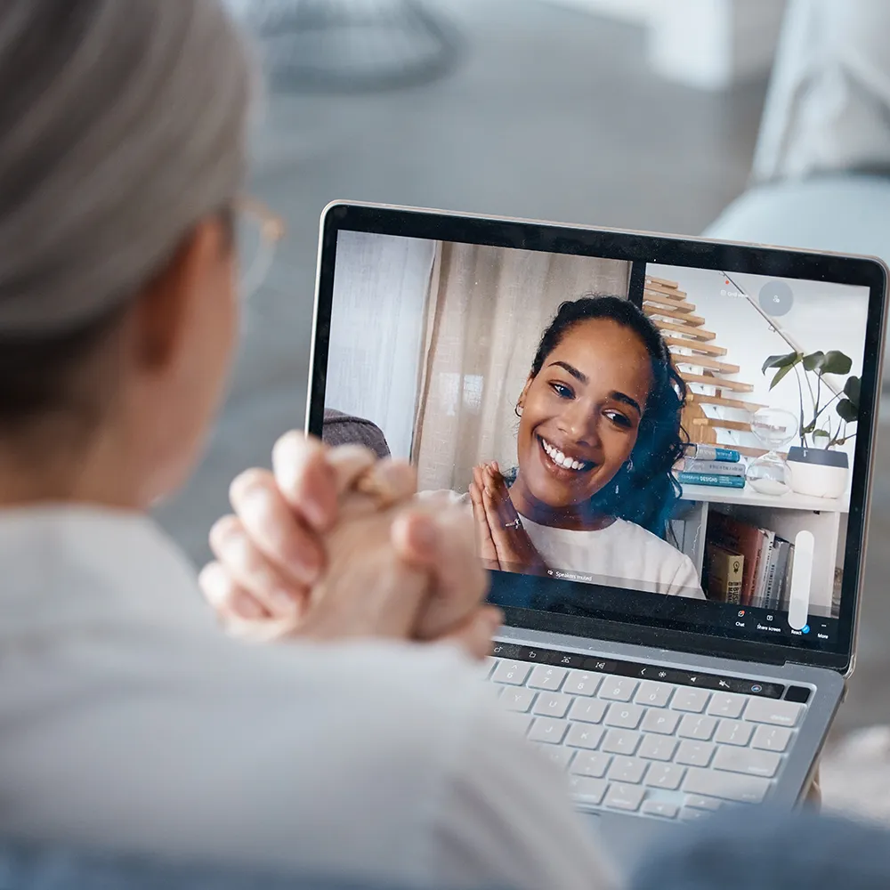 A person with gray hair sits with hands clasped, video chatting on a laptop with a smiling woman who appears on the screen; books and a potted plant are visible in the background.
