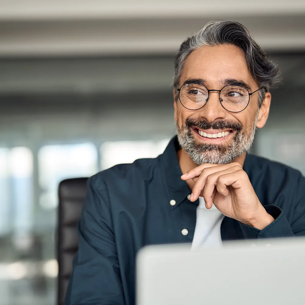A smiling man with glasses and salt-and-pepper hair sits at a desk, resting his chin on his hand, looking to the side in a modern office setting.
