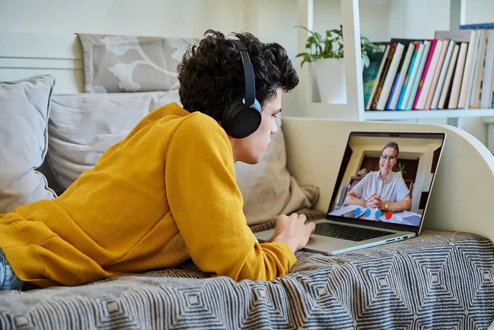 A person wearing headphones lies on a sofa, using a laptop for a video call with another person. Books and pillows are visible in the background.