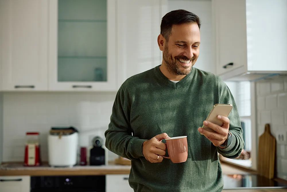 A man stands in a kitchen, smiling while looking at his smartphone. He is holding a pink mug in one hand and wearing a green sweater. The kitchen has white cabinets and wooden countertops.