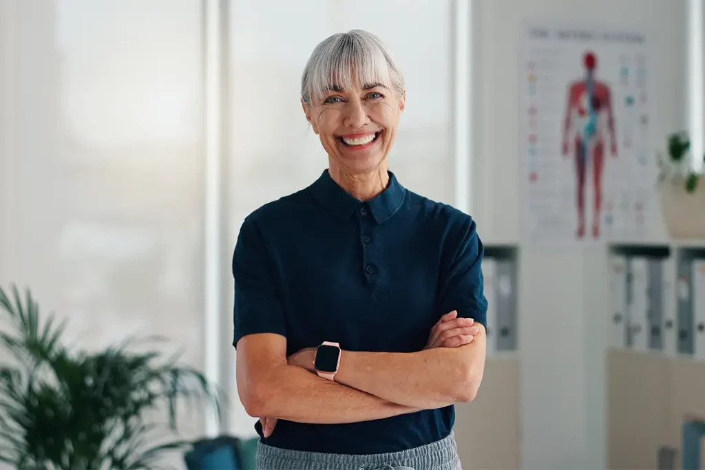 Smiling older woman with gray hair and bangs, wearing a navy blue polo shirt and a smartwatch, stands with arms crossed in a bright office with medical charts and plants in the background.