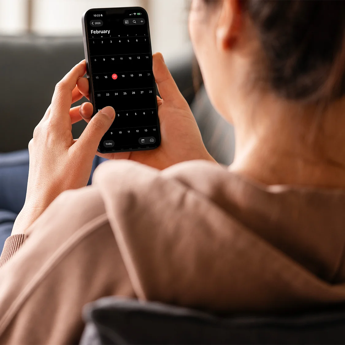 A person in a brown hoodie holds a smartphone, viewing a digital calendar set to February, with one date highlighted in red. The person is seated indoors, seen from behind and slightly over the shoulder.