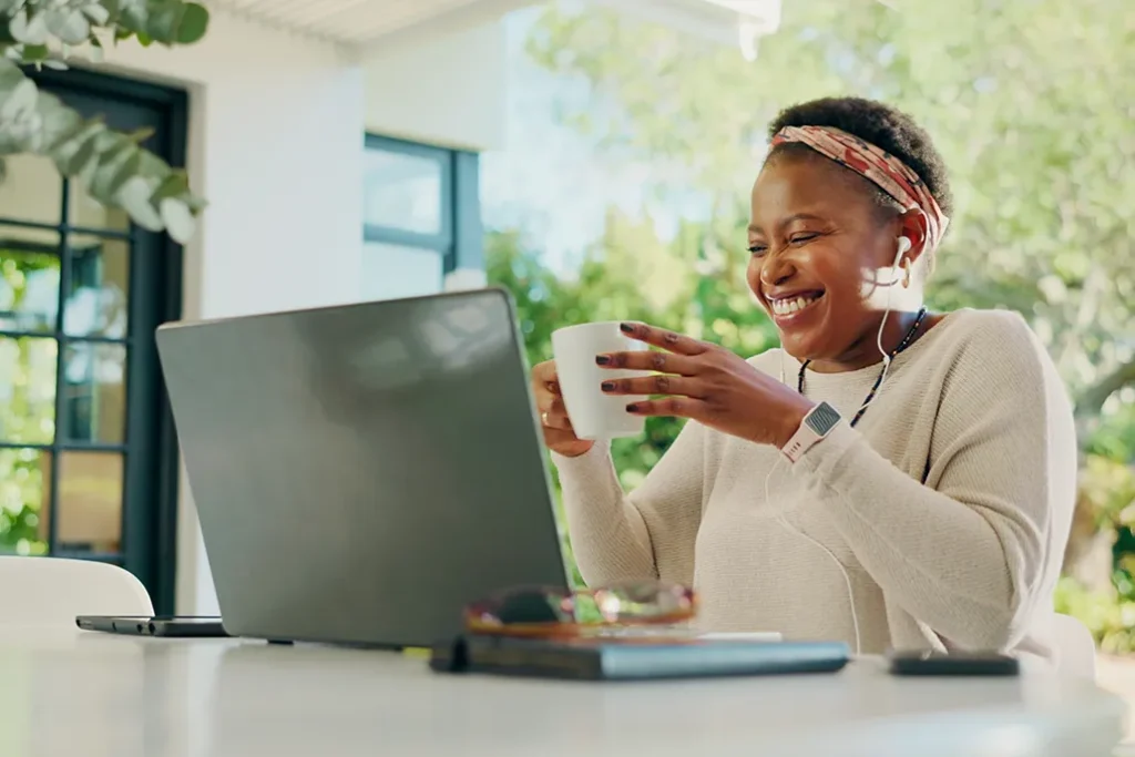 A woman smiles while holding a mug and looking at her laptop. She is wearing earphones and a headband, sitting at a bright table with notebooks and a smartphone, with greenery visible outside.