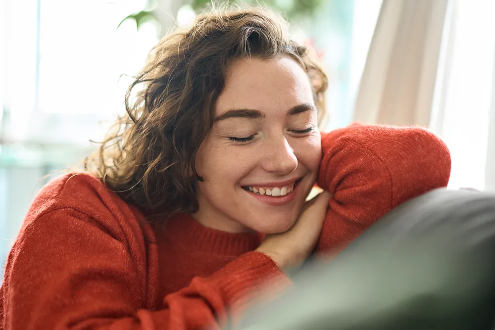 A person with curly brown hair wearing a red sweater smiles with their eyes closed, resting their head on their arms indoors near a window.