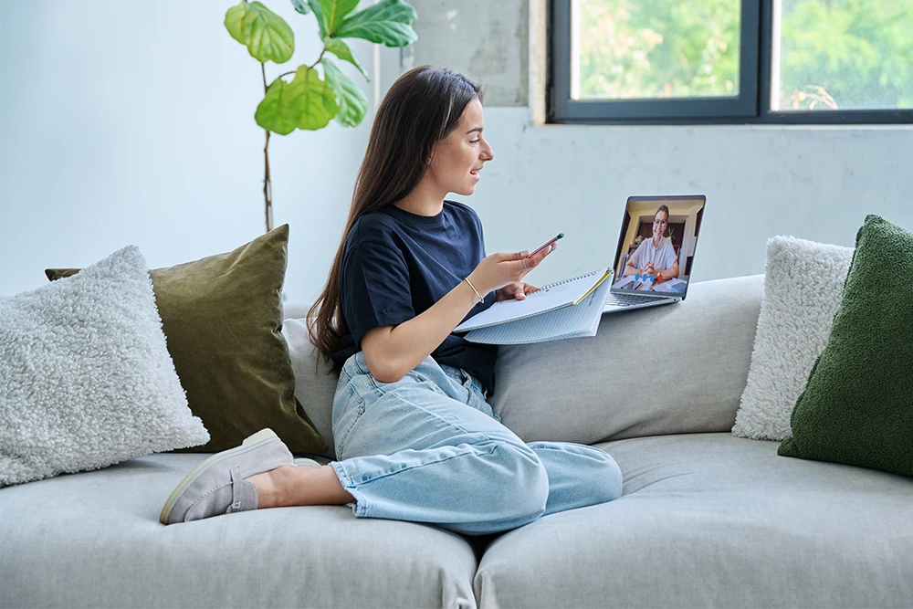 A young woman sits on a couch with notebooks, holding a phone and studying while having a video call on her laptop. She is engaged and relaxed, with a window and plant in the background.