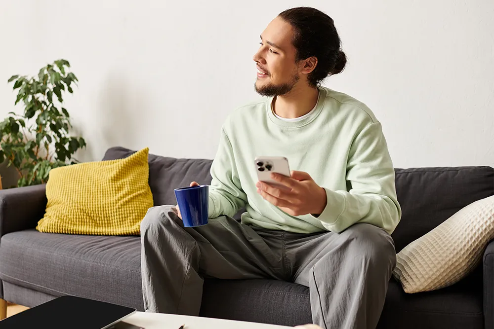 A man with a beard sits on a gray sofa, holding a smartphone in one hand and a blue mug in the other. He is wearing a light green sweatshirt and gray pants, with yellow and beige cushions beside him.