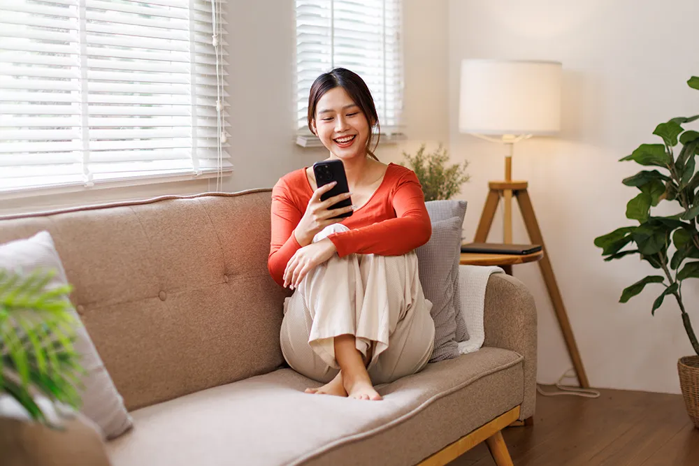 A woman sits barefoot on a beige sofa, smiling while looking at her phone. She wears a red long-sleeve top and light pants. There are plants, a lamp, and sunlight coming through window blinds in the cozy living room.
