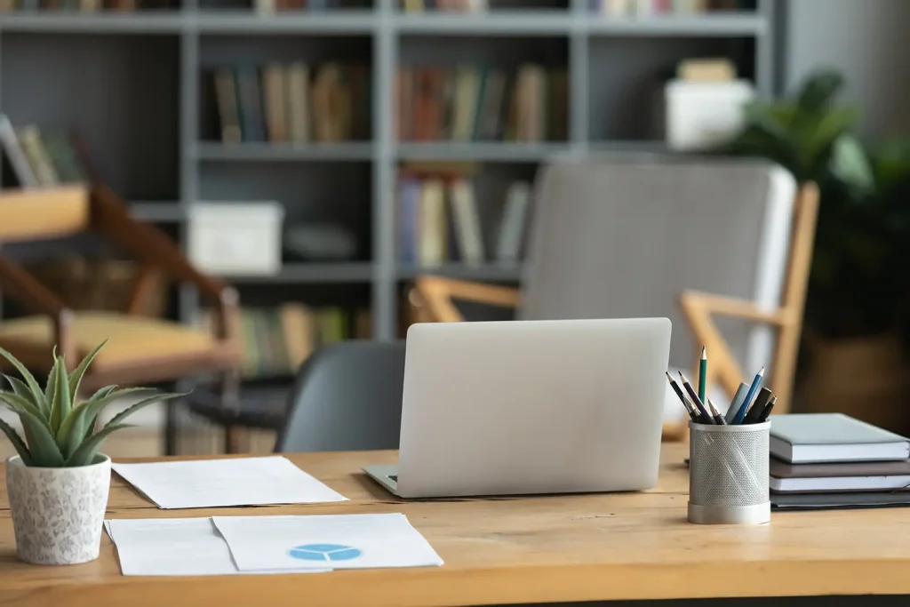 A tidy desk with a laptop, papers, a potted plant, pens in a holder, and notebooks, set in a cozy office with bookshelves and chairs in the background.