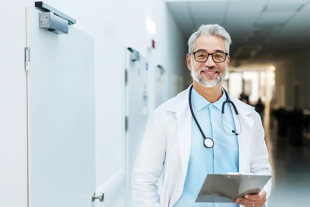 A smiling doctor with gray hair and glasses stands in a bright hallway, wearing a white coat and stethoscope, holding a clipboard. The mood is professional and welcoming.