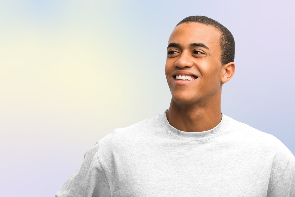 A young man wearing a light gray sweatshirt smiles and looks to his right, standing against a soft gradient pastel background.