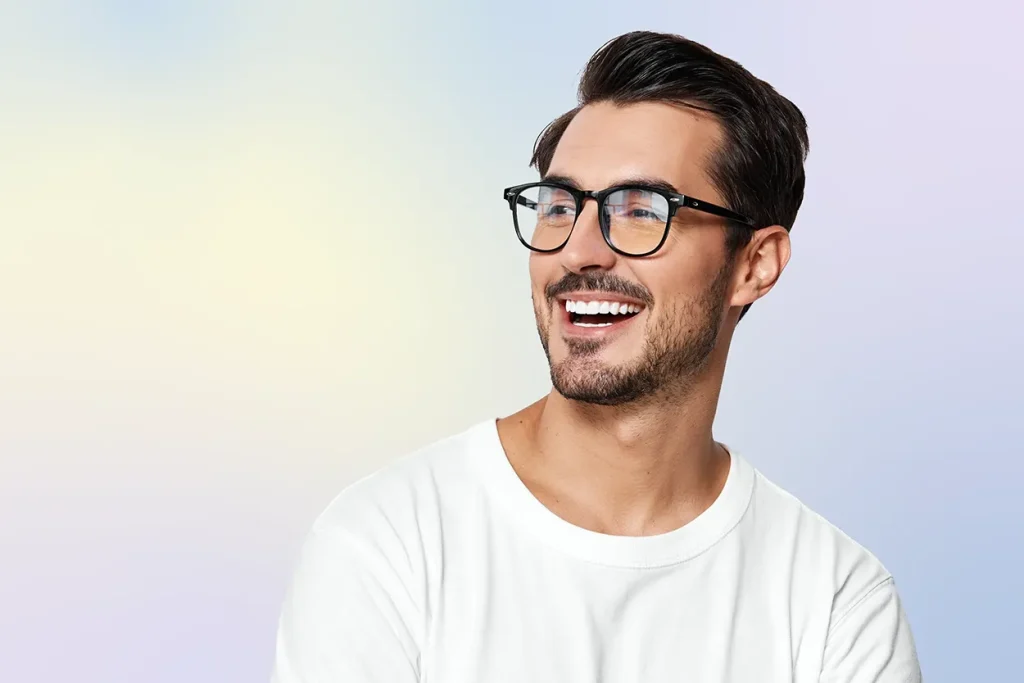 Smiling man with glasses and a white shirt against a soft pastel gradient background. He looks to the side, conveying a cheerful and relaxed mood.